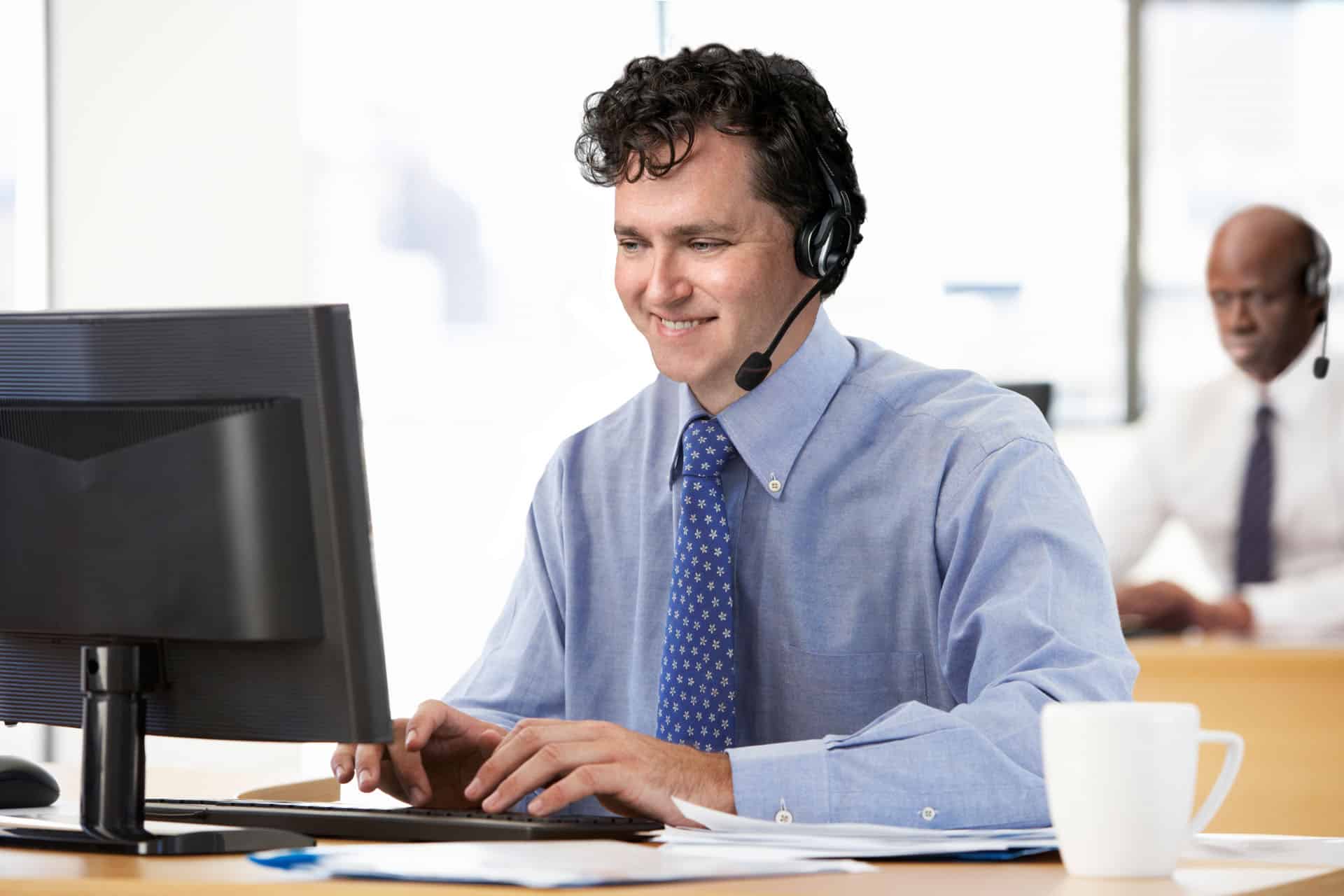A man wearing a headset sits at a desk, typing on a computer keyboard. He is smiling, embodying the collaborative spirit of an explainer video company. Another person in the background is also working at a desk.