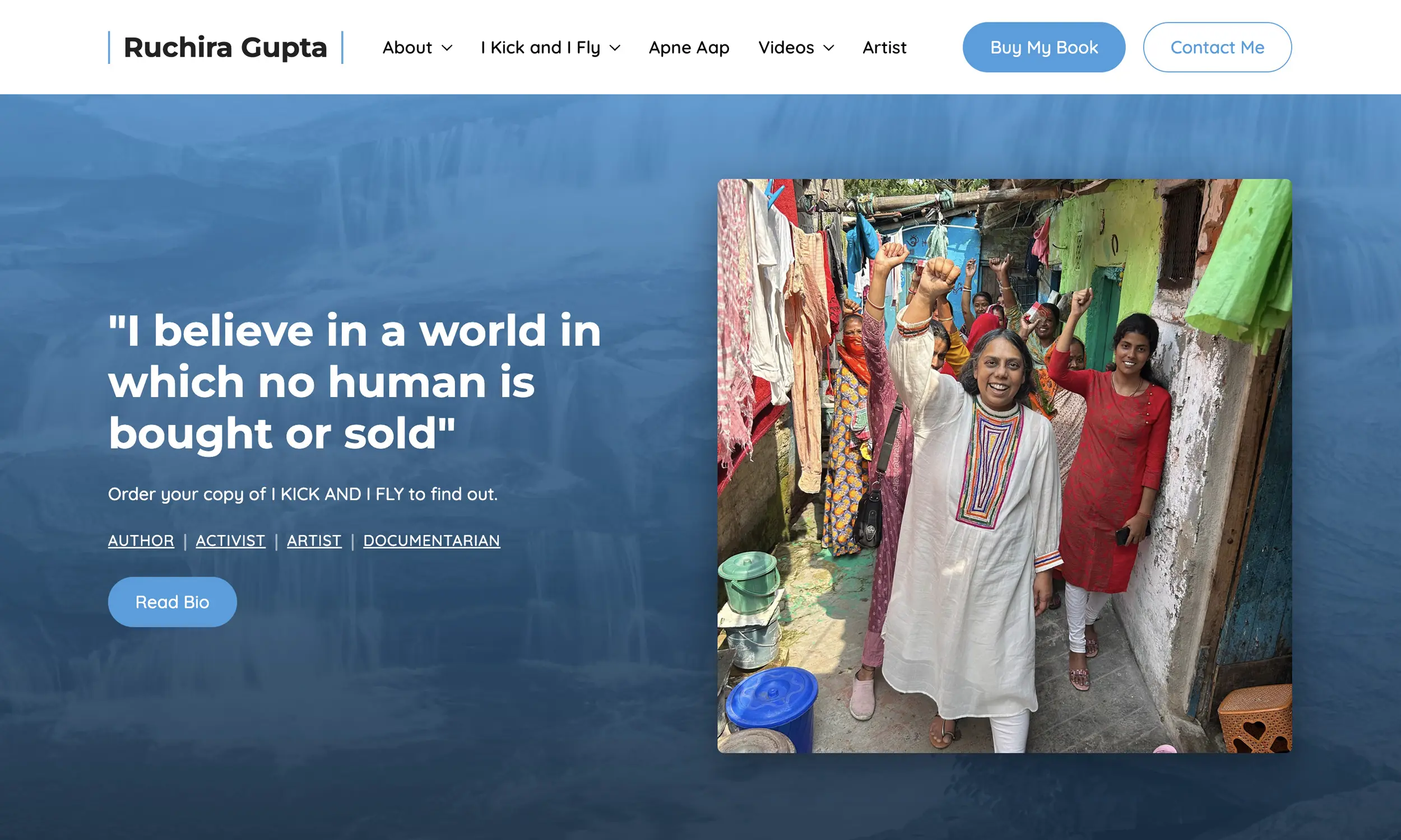 A woman in a white kurta raises her fist and smiles while walking through a narrow alley, as if celebrating an author website redesign, with other women and laundry hanging in the background.
