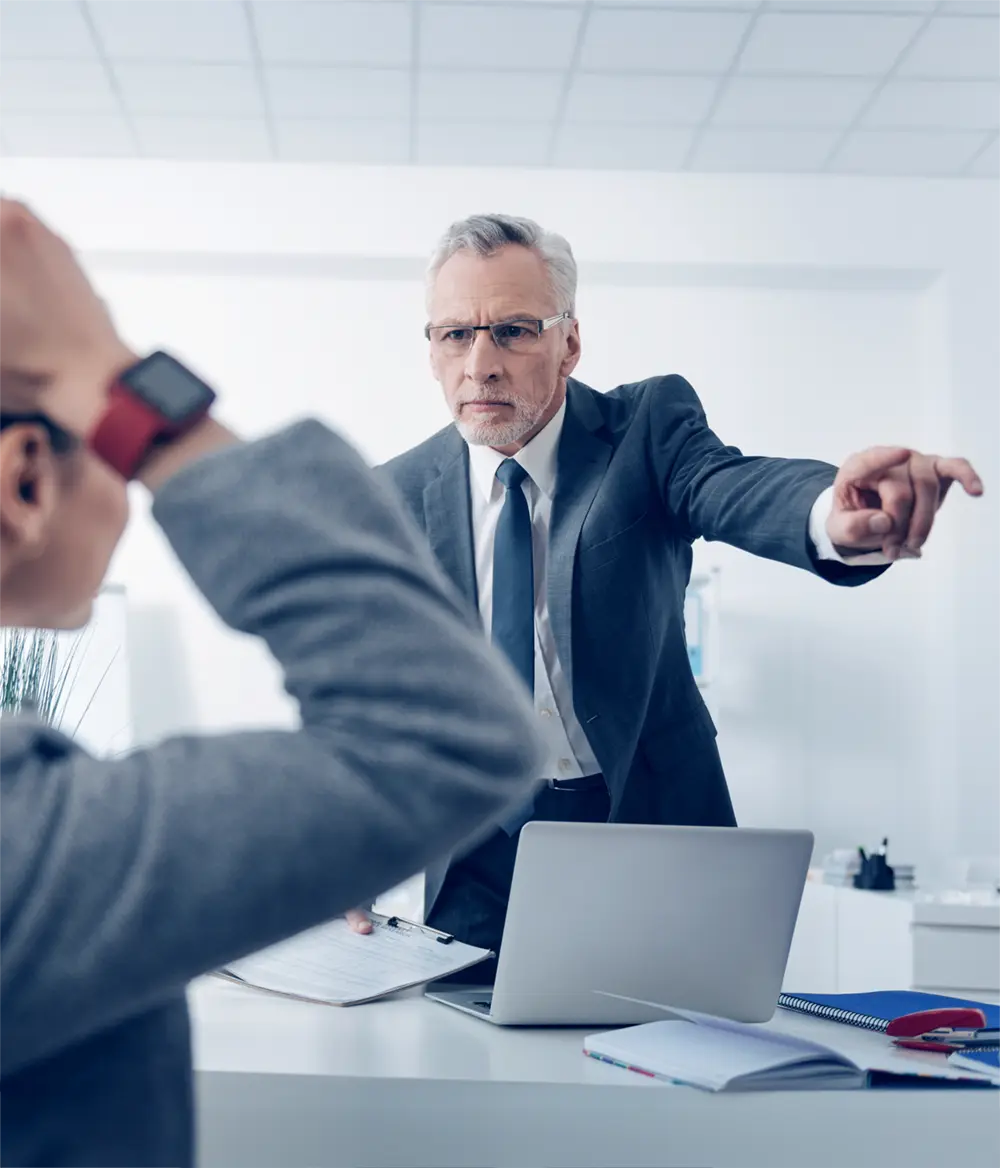 A man in a suit points angrily at a seated person in an office setting, while the seated person holds their head in apparent distress.