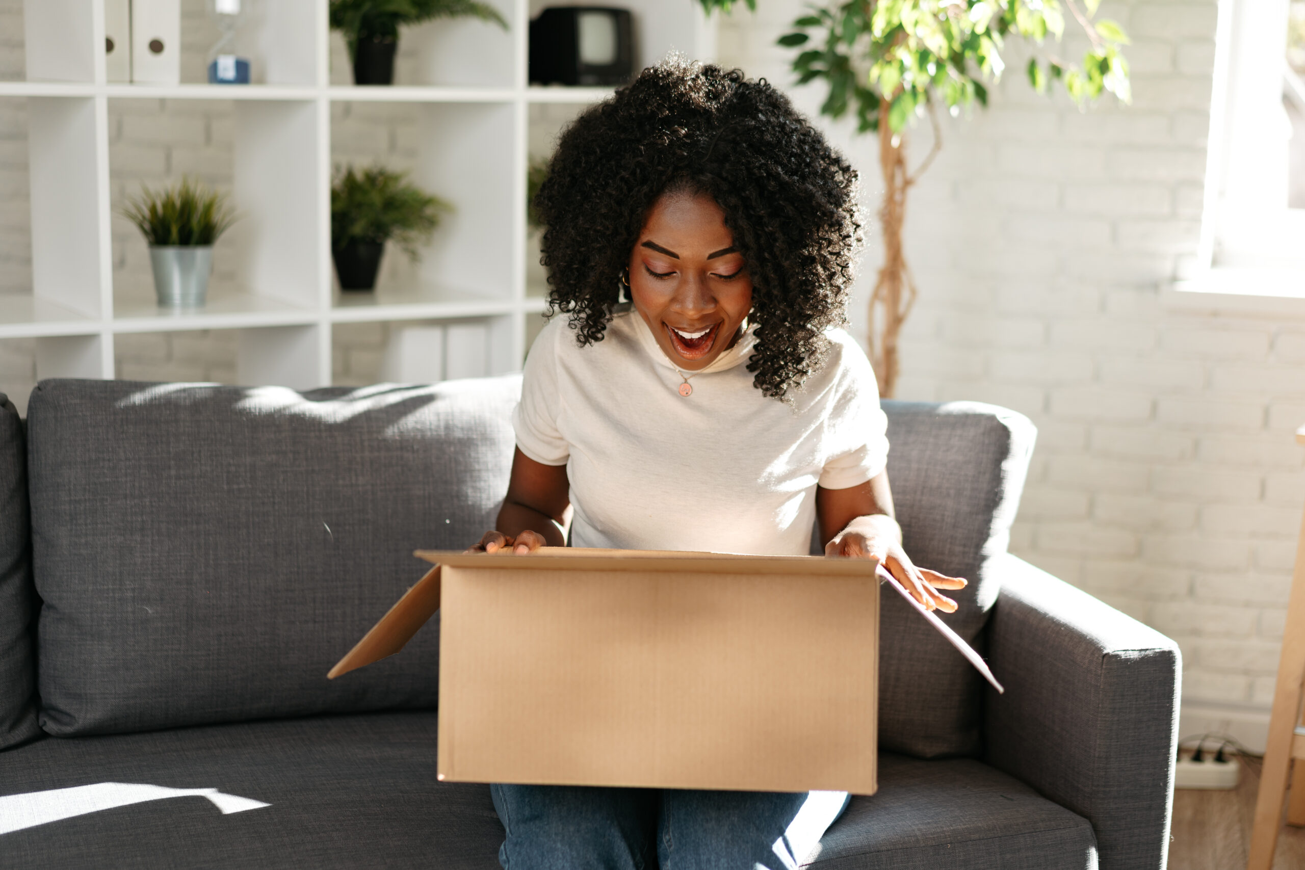 A woman sitting on a couch opens a cardboard box, looking inside with a surprised and happy expression. Shelves with plants and decor are visible in the background.