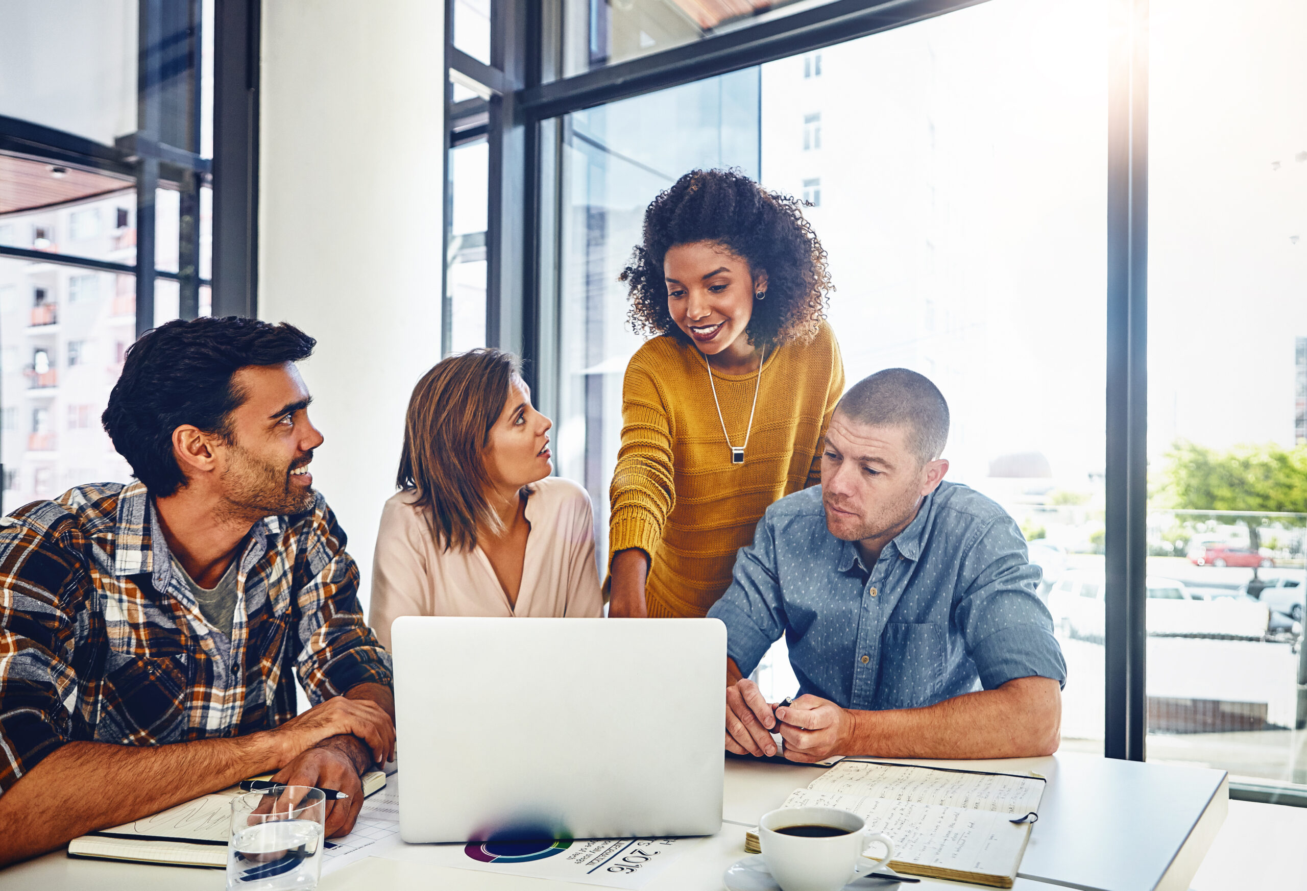 Four people sit around a table with a laptop, notebooks, and coffee, engaged in discussion about website design cost in a bright office setting with large windows.