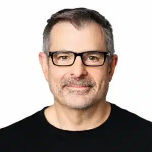 Middle-aged man with short gray hair, trimmed beard, and glasses, wearing a black shirt, facing the camera against a plain white background.