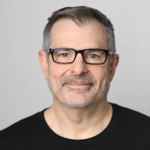 A middle-aged man with short gray hair, glasses, and facial stubble, wearing a black crew-neck shirt, smiles slightly against a plain light gray background.