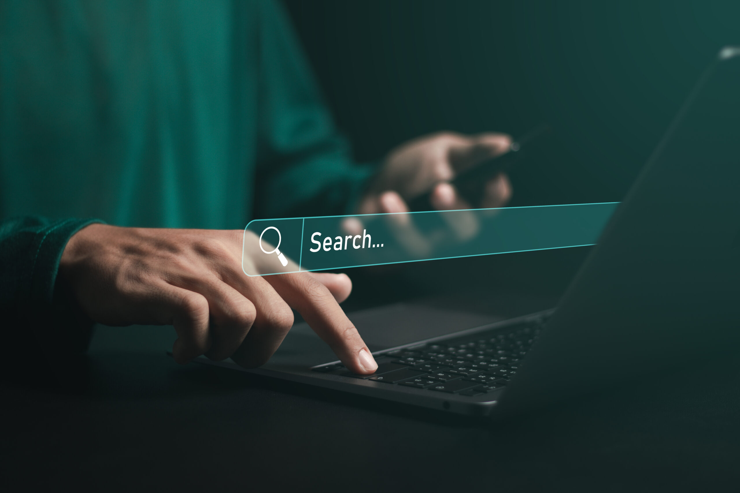 Person using a laptop and smartphone with a virtual search bar overlayed on the image, suggesting internet browsing or comparing SEO vs PPC strategies for online searching.