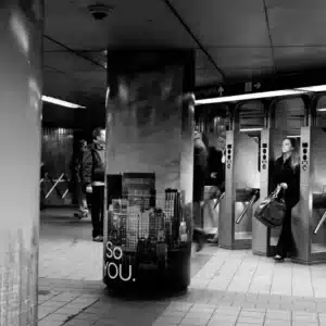 Black-and-white photo of people in a subway station; a pillar with a cityscape and the words "SO YOU." is in the foreground. Turnstiles and a sign reading "To North" are visible.