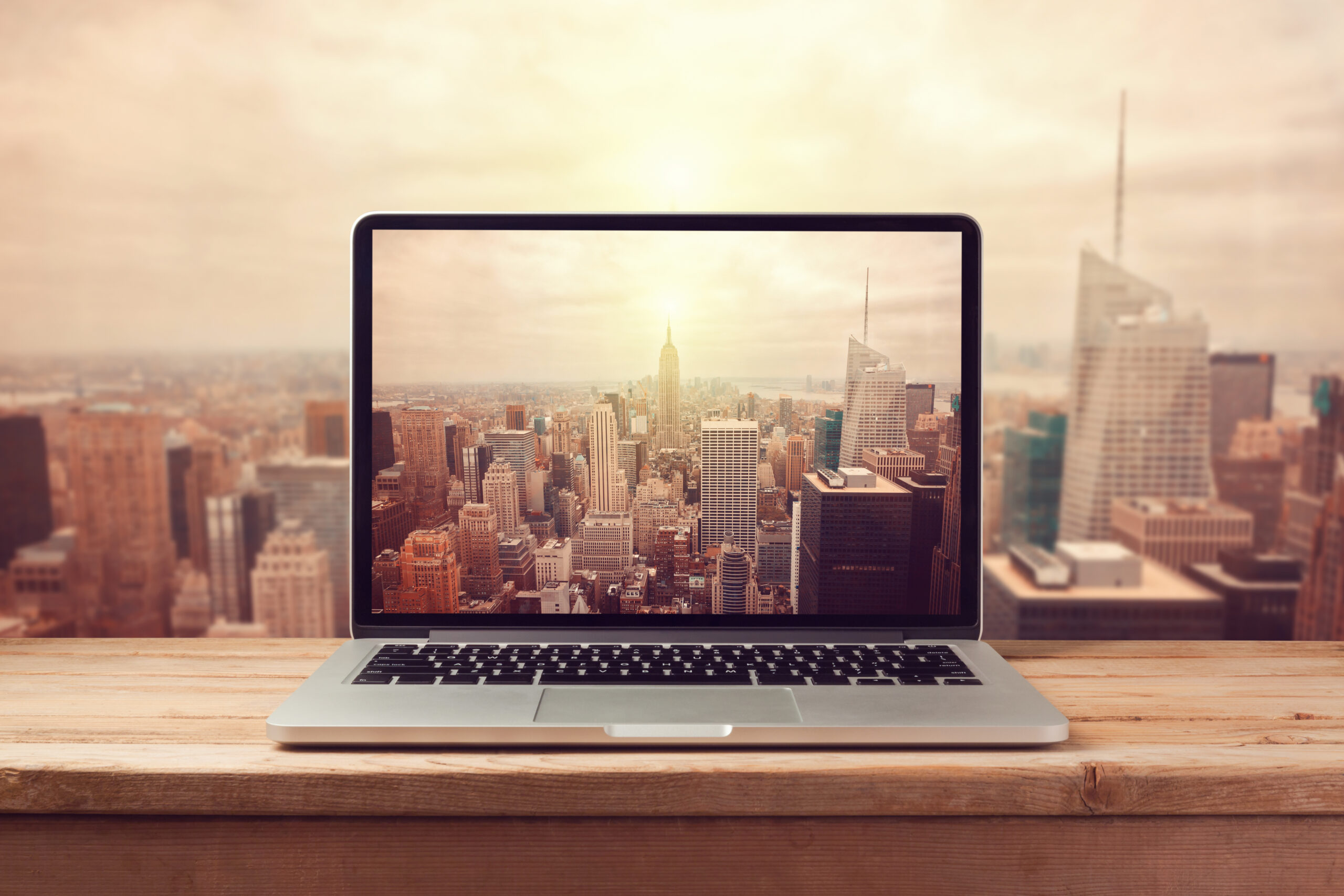 A laptop on a wooden table displays a city skyline on its screen, with a similar city view seen in the background, suggesting a blend of virtual and real perspectives.