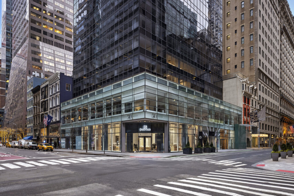 Glass-walled modern building on a city corner, surrounded by high-rise buildings, with a few parked cars and empty sidewalks.