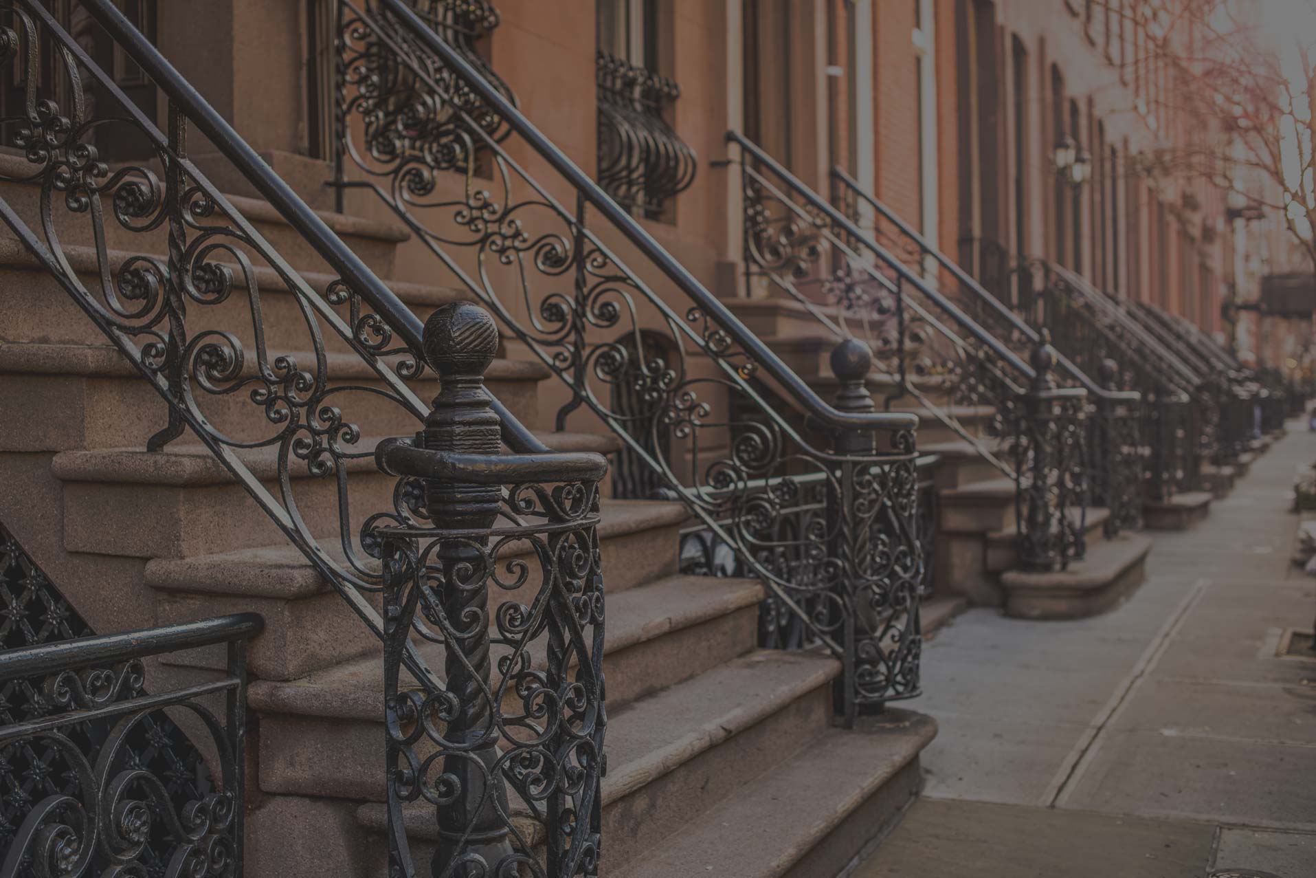 The main picture on LC|NYC. A row of brownstone stoops with ornate wrought iron railings lines a city sidewalk.