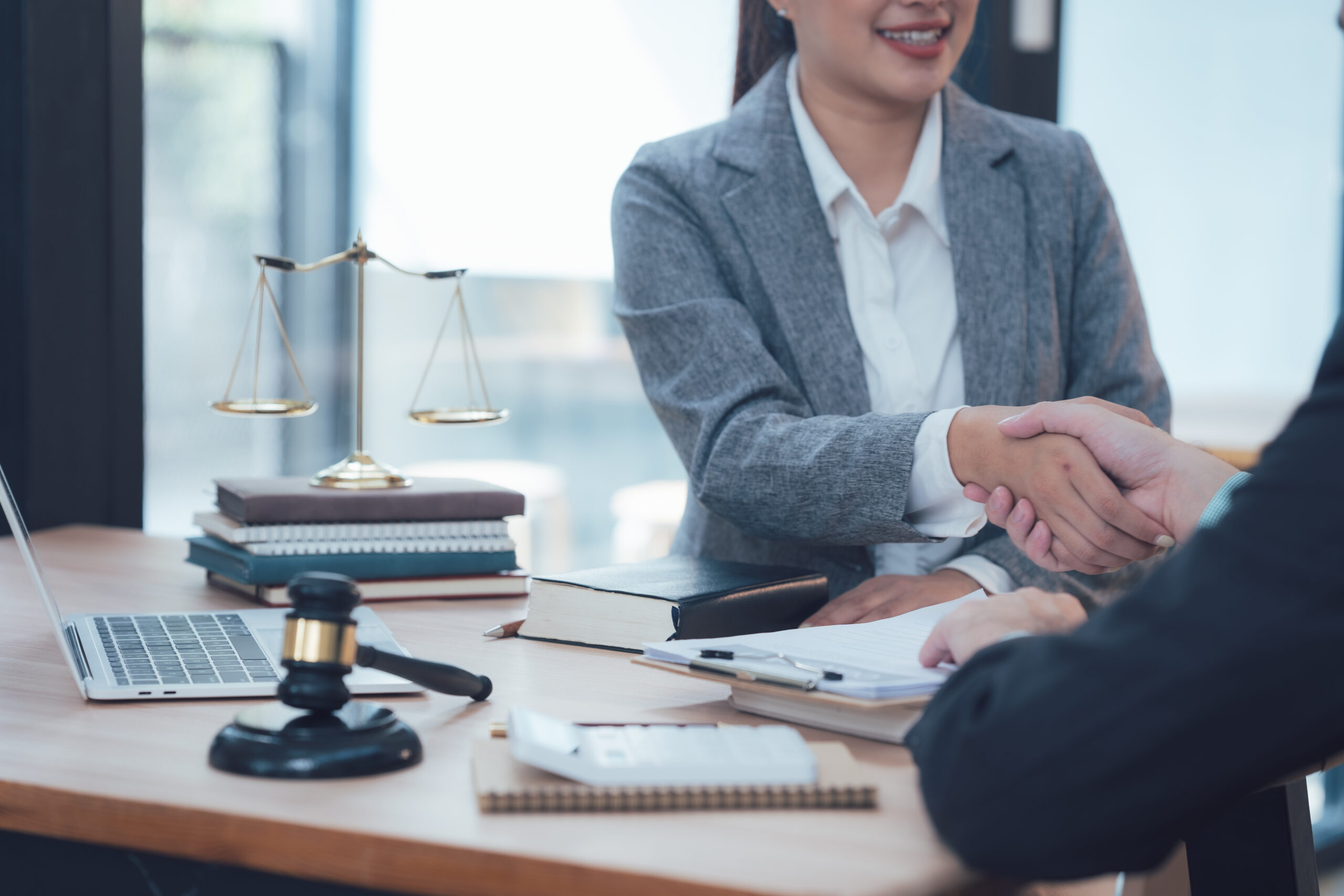 Two people in business attire shake hands at a desk with legal books, a gavel, scales of justice, and a laptop—highlighting the importance of Law Firm SEO in today’s office setting.