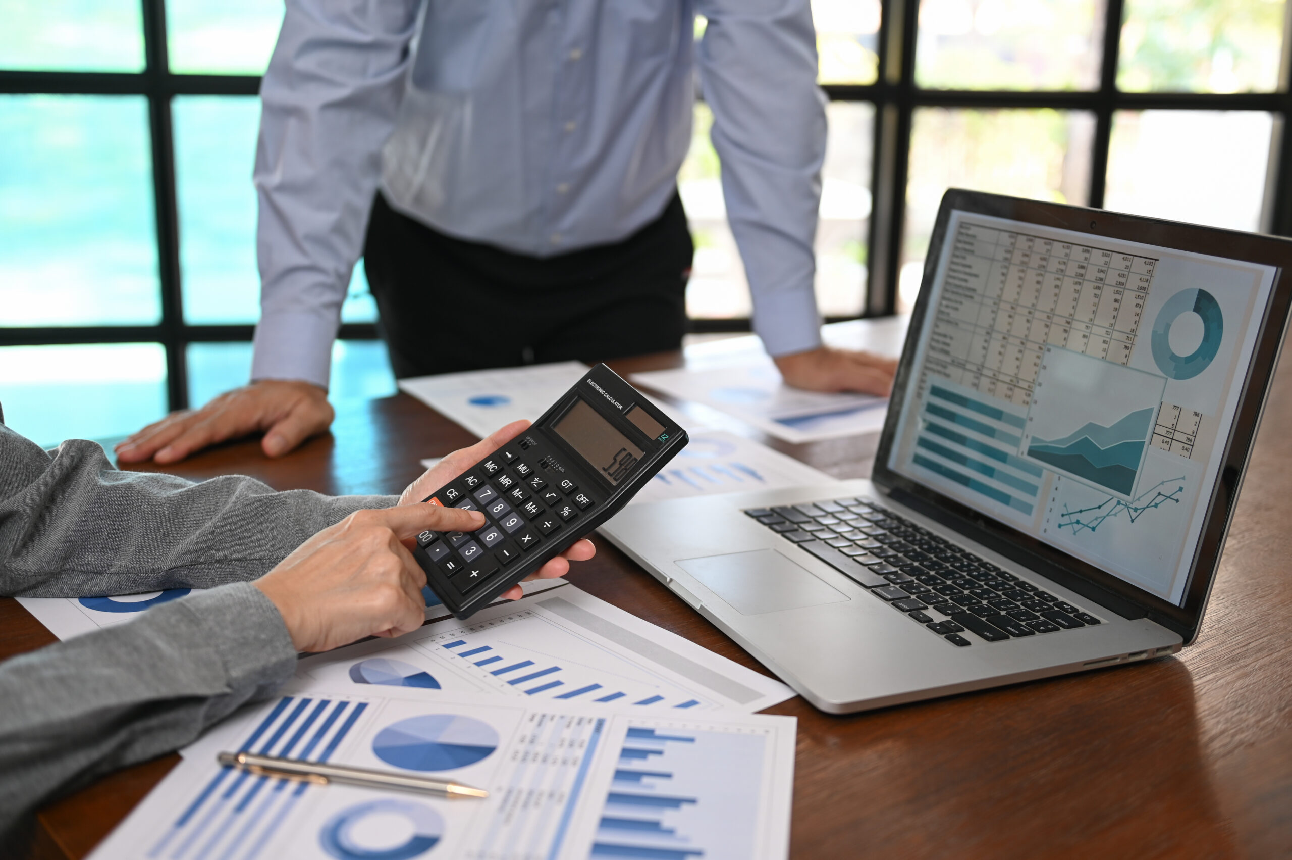 Two people reviewing financial charts and graphs on paper and a laptop, with one person using a calculator at a wooden desk—analyzing data critical for SEO For Accountants success.