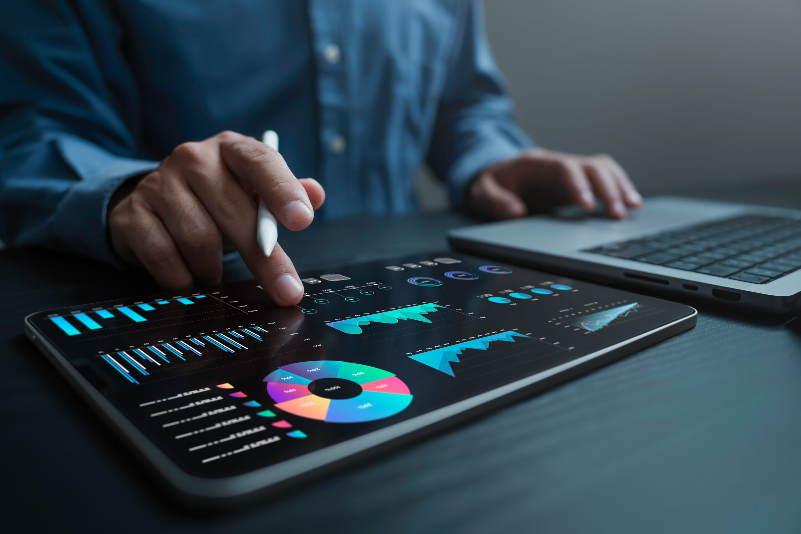 A person uses a stylus to interact with financial charts and graphs displayed on a tablet, with a laptop nearby on a dark desk.