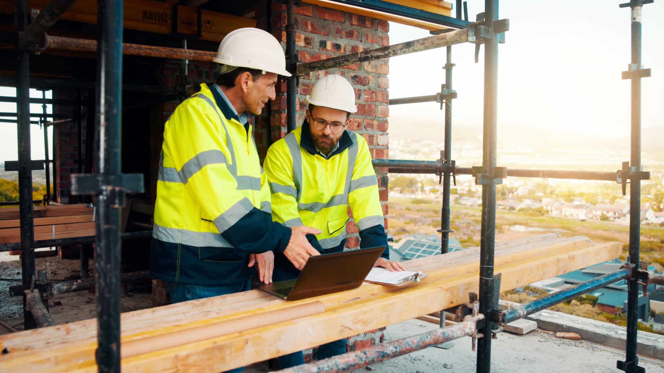 Two construction workers in safety gear and helmets review plans on a laptop at a building site, surrounded by scaffolding and construction materials—demonstrating how technology like SEO for Contractors can streamline project planning and enhance efficiency on the job.