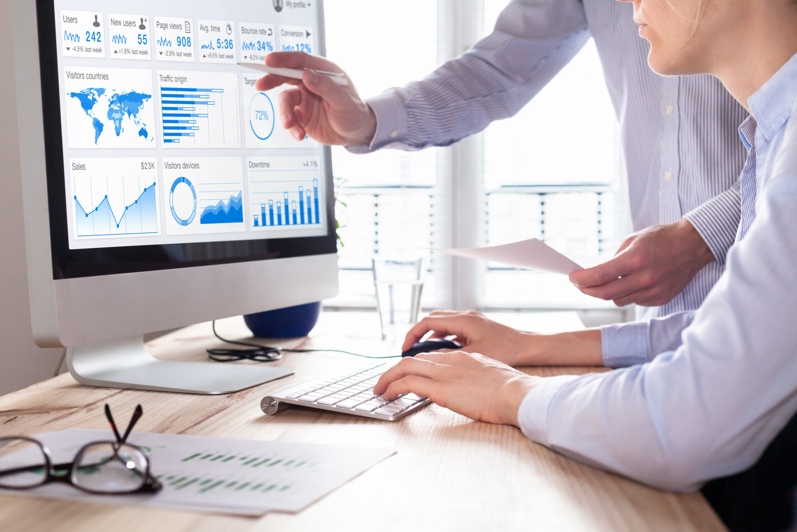 Two people in an office setting analyze data and charts on a computer monitor, reviewing graphs, percentages, and reports—ideal for Web Design Firms seeking insights to boost digital strategy.