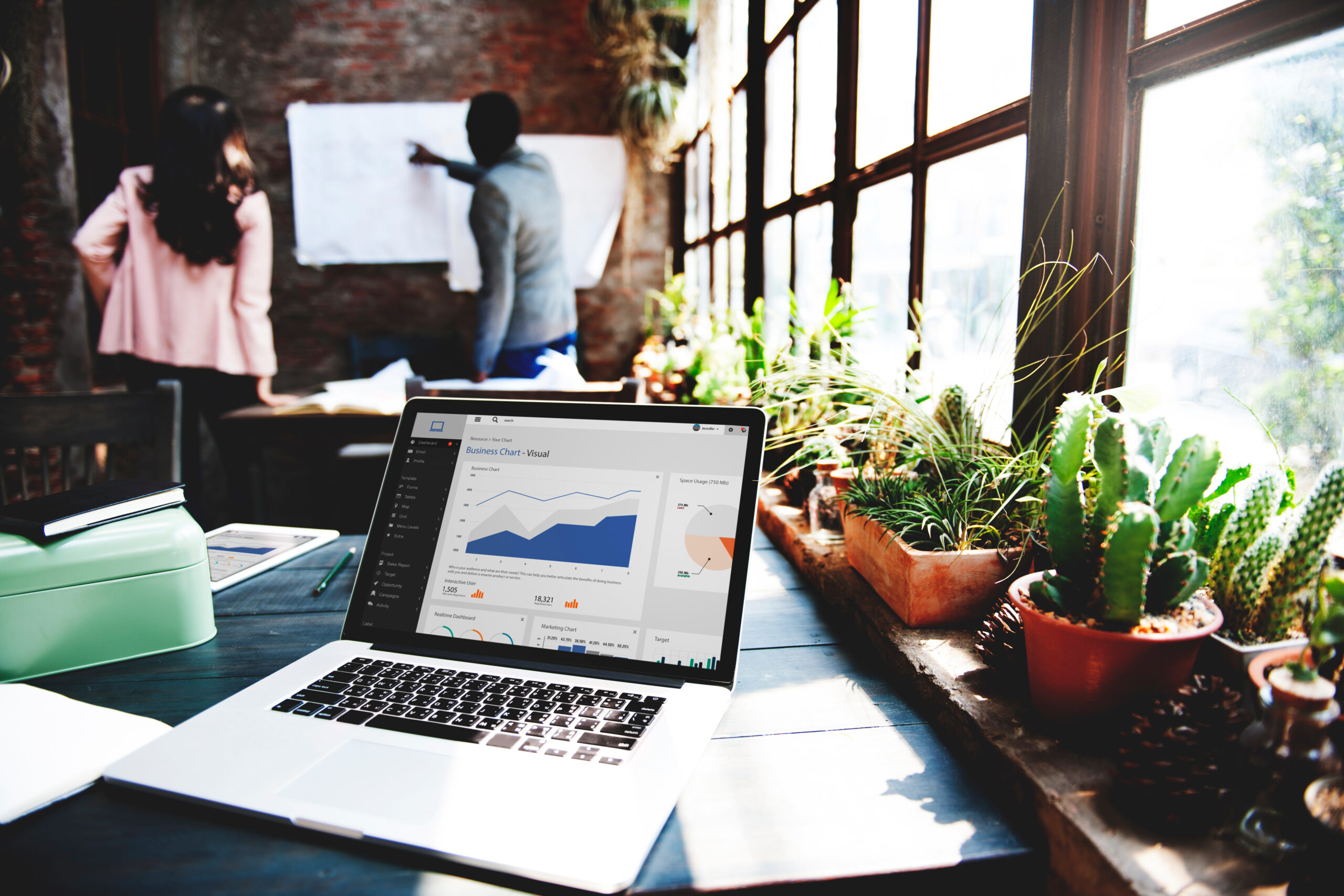 Open laptop displaying business analytics on a table with potted plants; two people from the Best Marketing Agency discuss a chart on a whiteboard in the background.