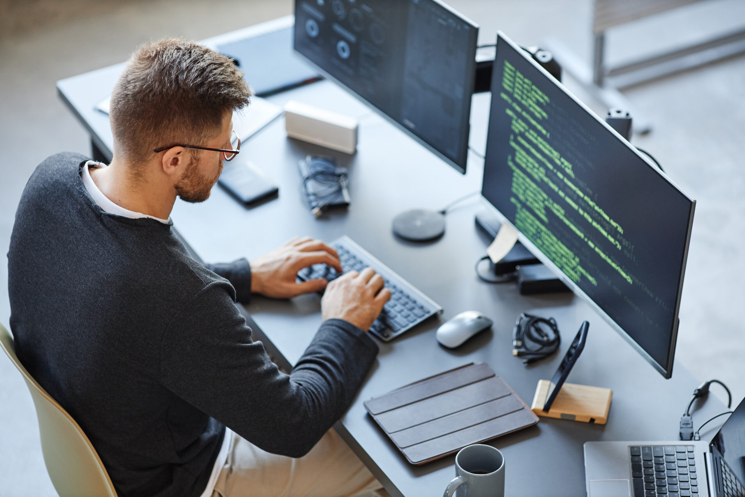 Man working at a desk with two monitors displaying code, typing on a keyboard. Various office items and electronics are on the desk.