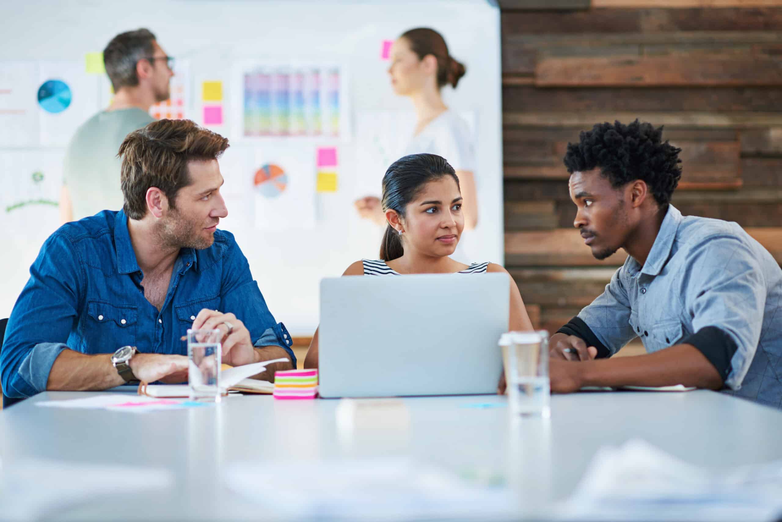 Three people sit at a table with a laptop, discussing work, while two others stand near a whiteboard with charts and sticky notes in the background.