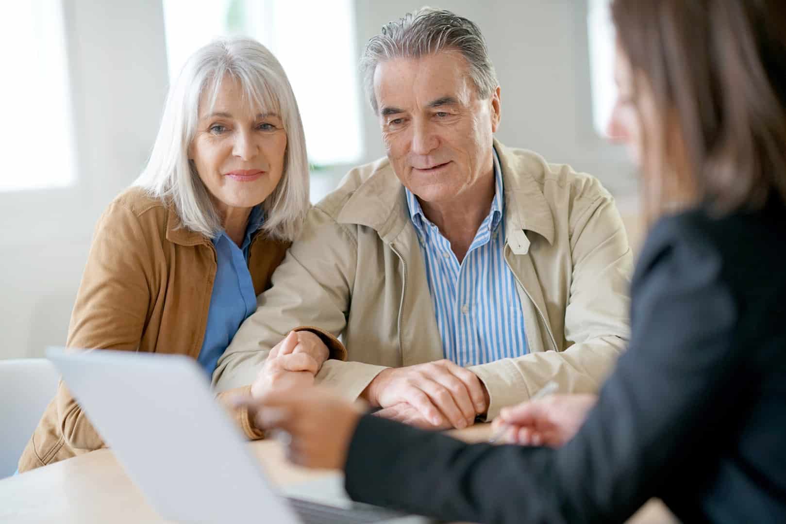 An older couple sits at a desk, looking at a laptop screen while a woman in business attire points and explains something to them.