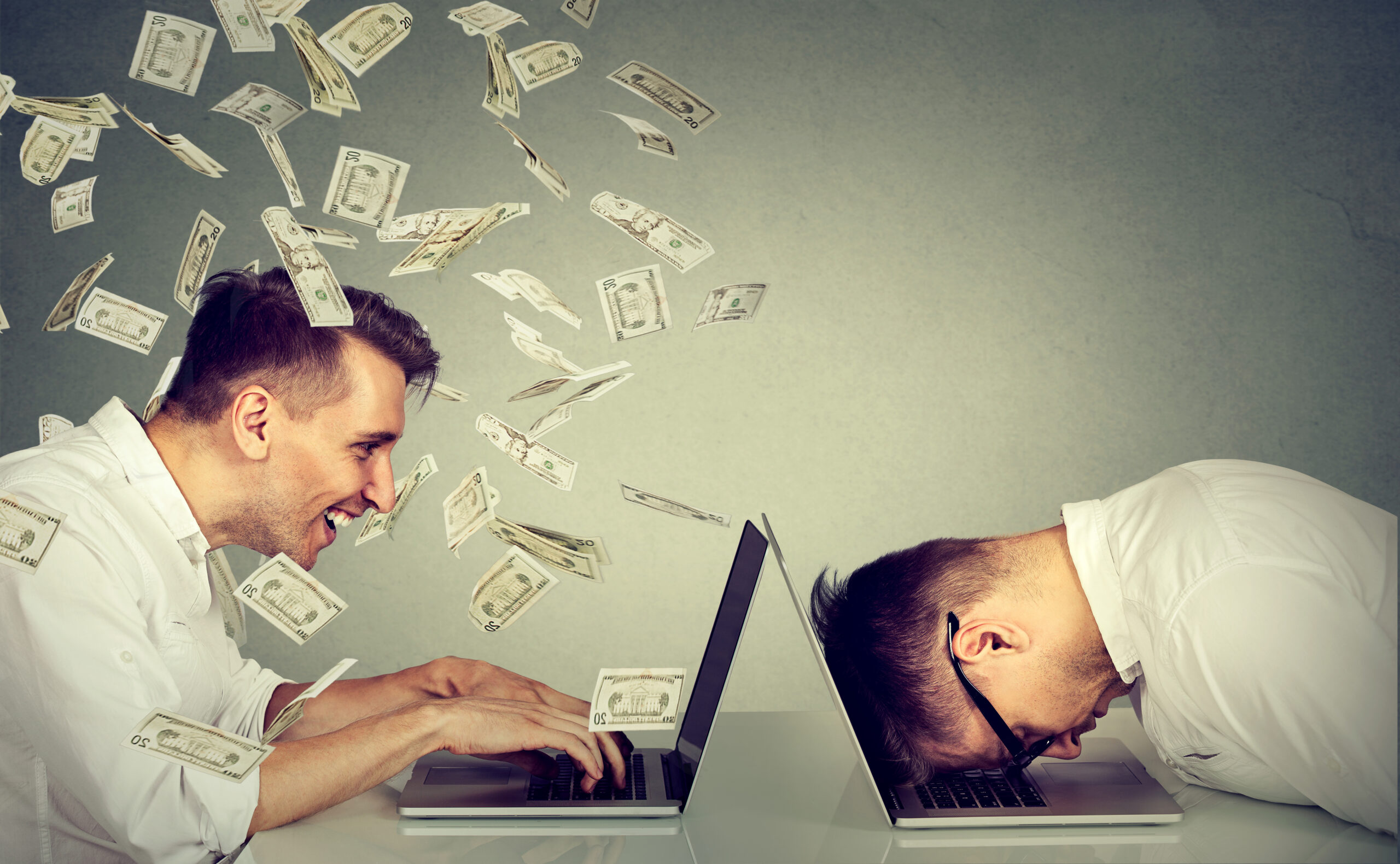 Two men sit at laptops; one smiles as money flies from his screen, while the other has his head down in frustration as money falls away.