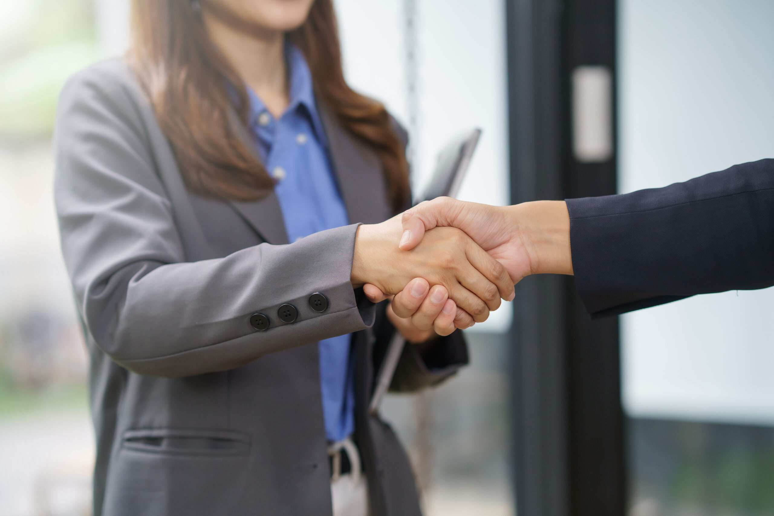 Two people in business attire shake hands indoors, one holding a tablet, suggesting a professional agreement or meeting.