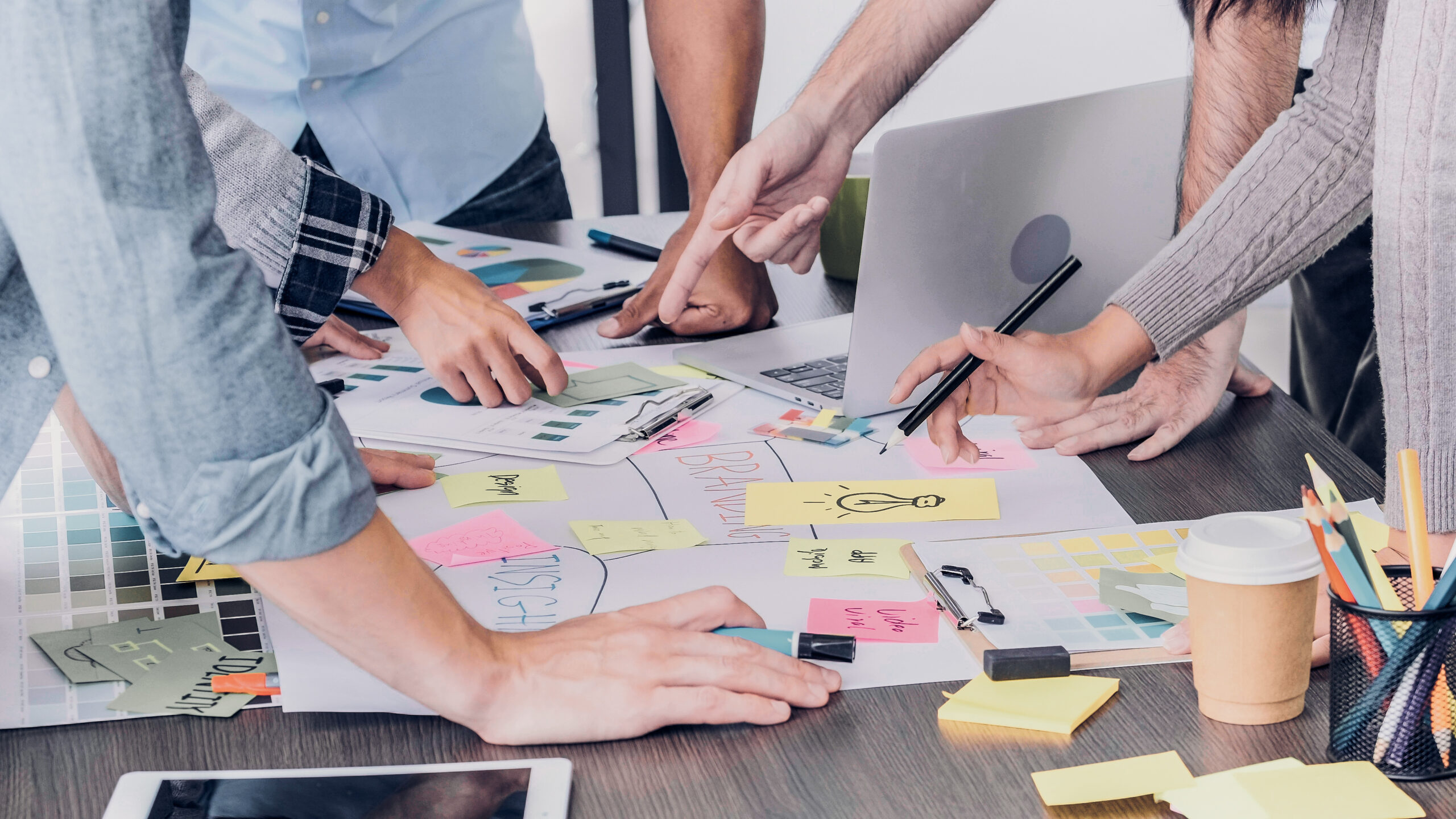 People working together at a table covered with papers, sticky notes, a laptop, color swatches, and stationery, appearing to collaborate on a project.