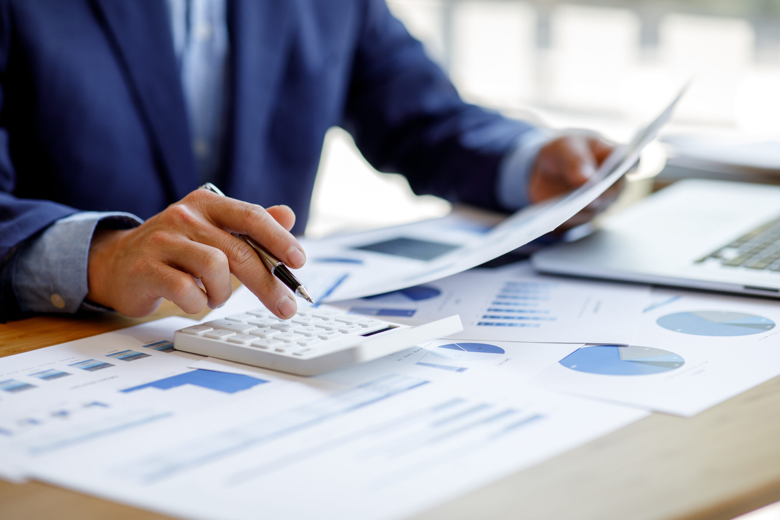 Person in business attire uses a calculator and reviews financial documents with charts and graphs at a desk with a laptop.