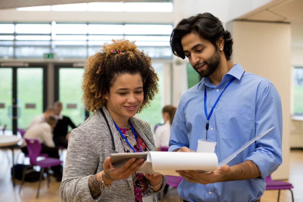 Two professionals with ID badges review documents together in a bright office space, with people collaborating in the background.