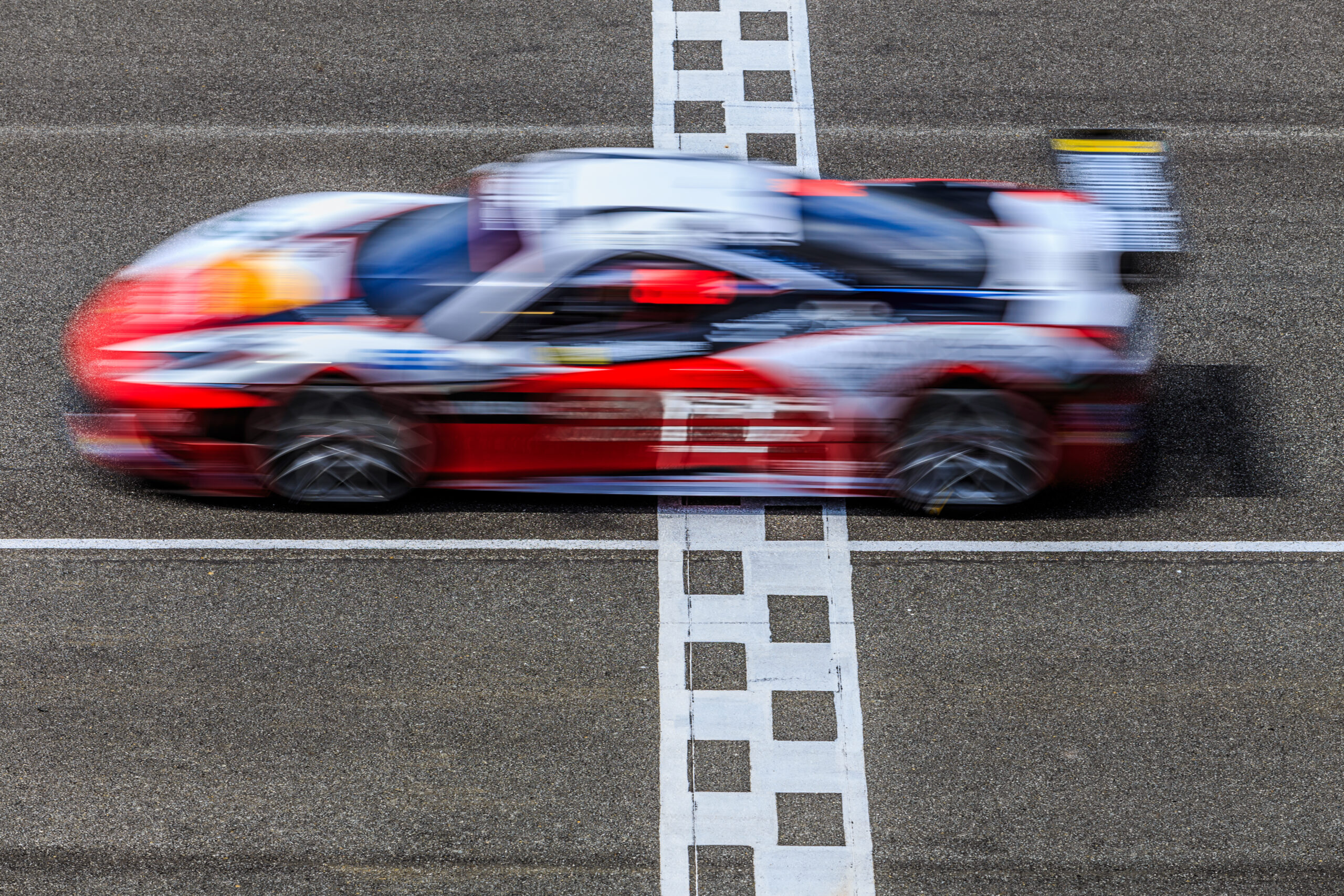 A red and white race car crosses a checkered finish line on a track, captured in motion blur.