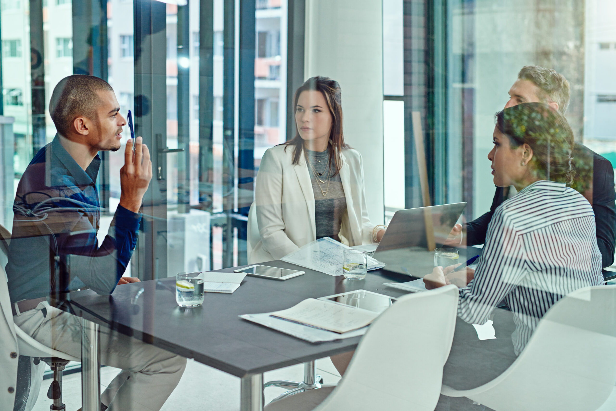 Four people sit around a conference table in an office, engaged in discussion with laptops, papers, and glasses of water in front of them.