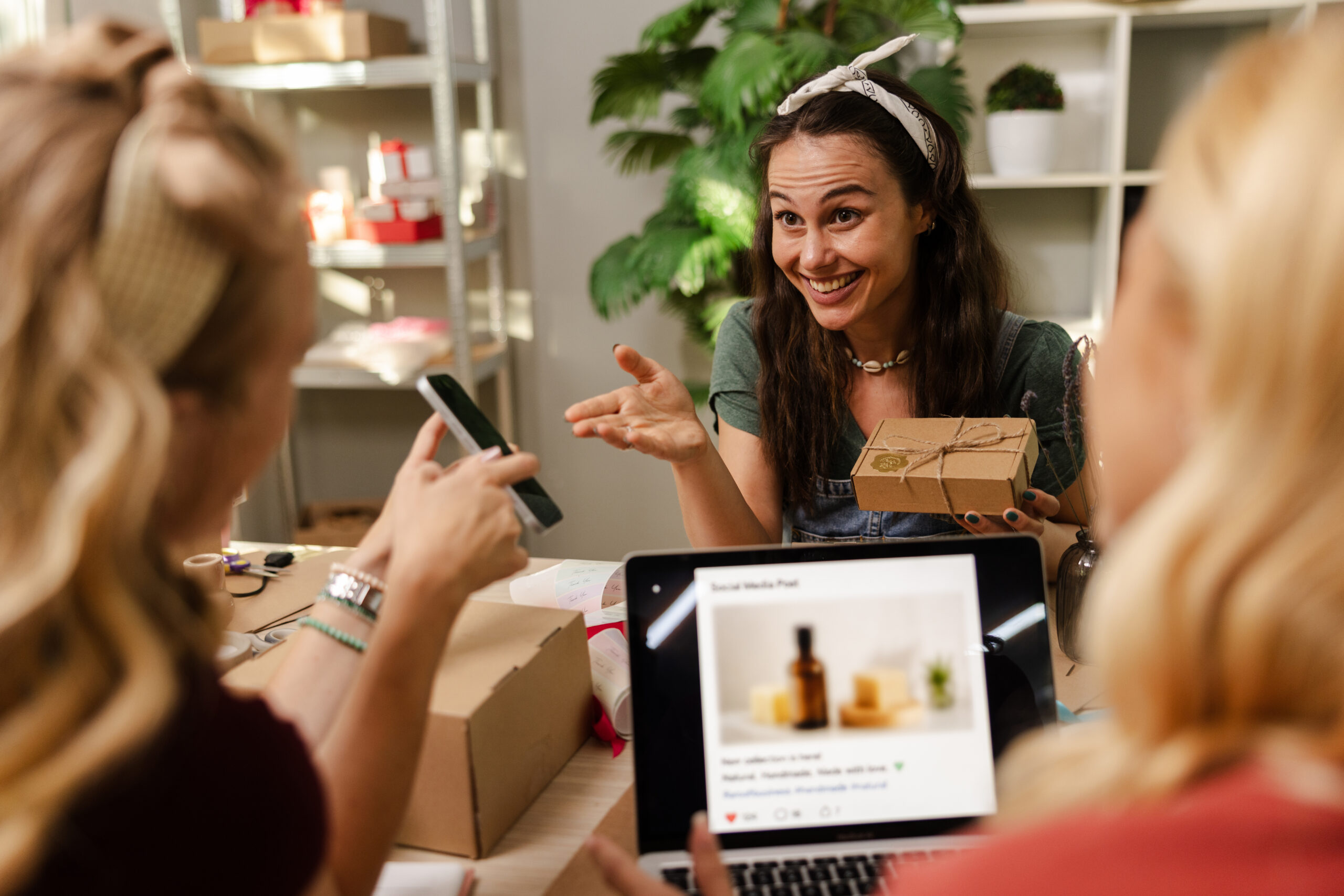 Two women sit at a table; one smiles while holding a gift box, and a laptop showing a social media post is open in front of them.