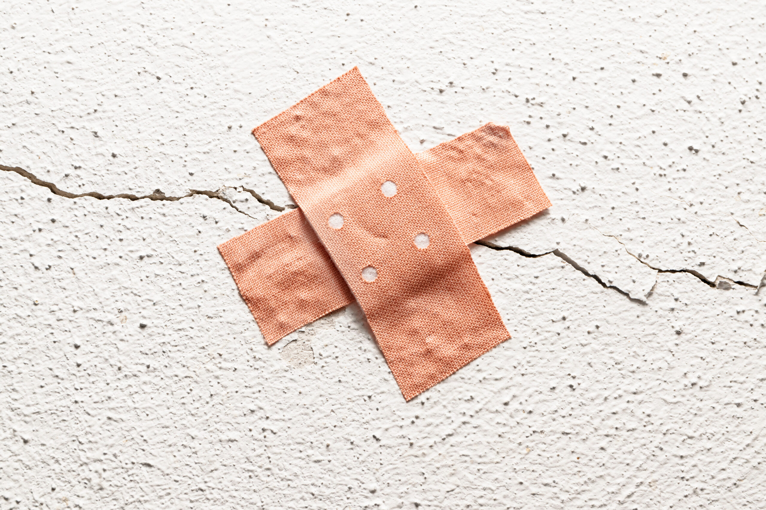 Two adhesive bandages arranged in an X shape are placed over a crack on a textured white wall.