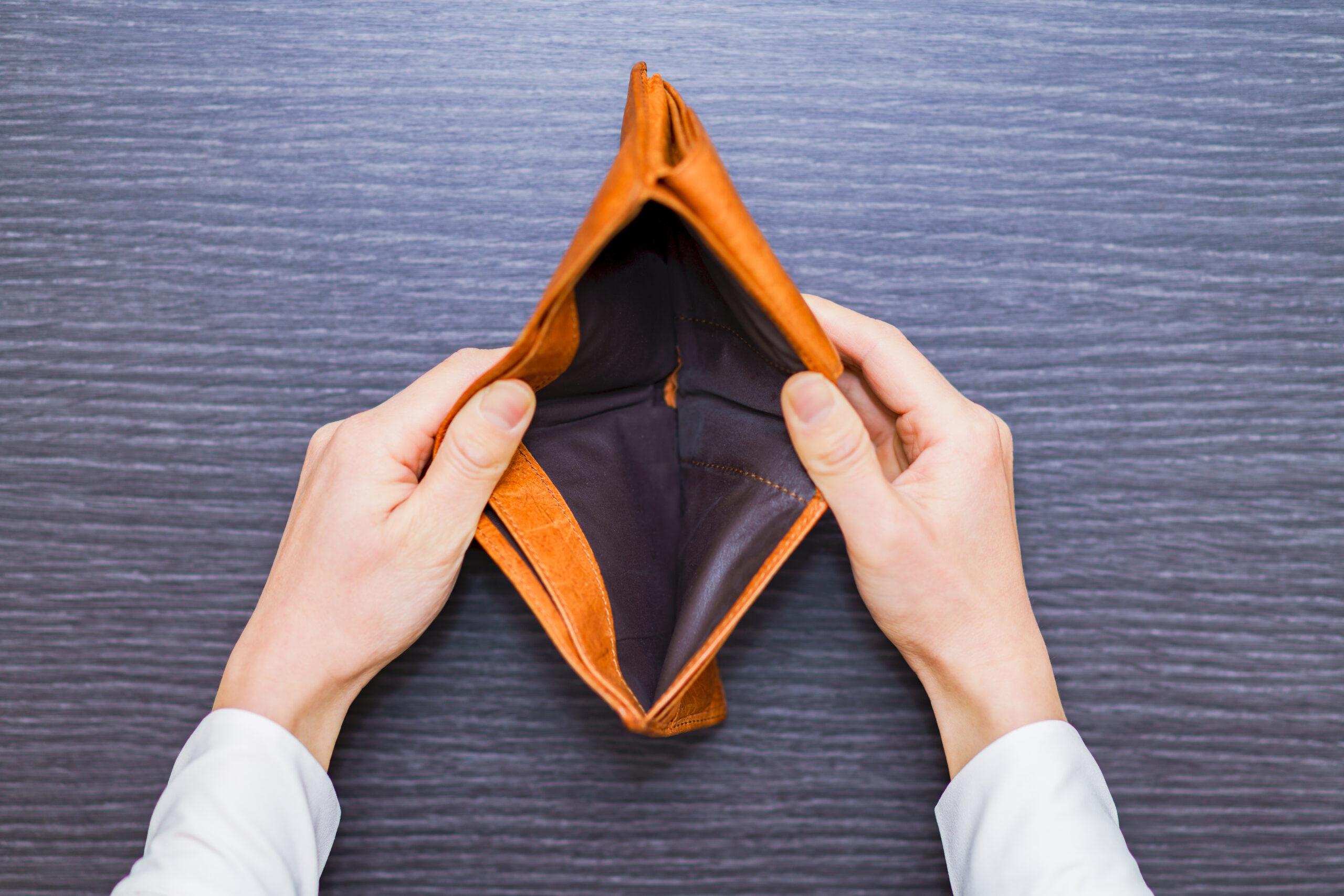 A person holds open an empty brown wallet over a dark wooden surface, showing there is nothing inside.