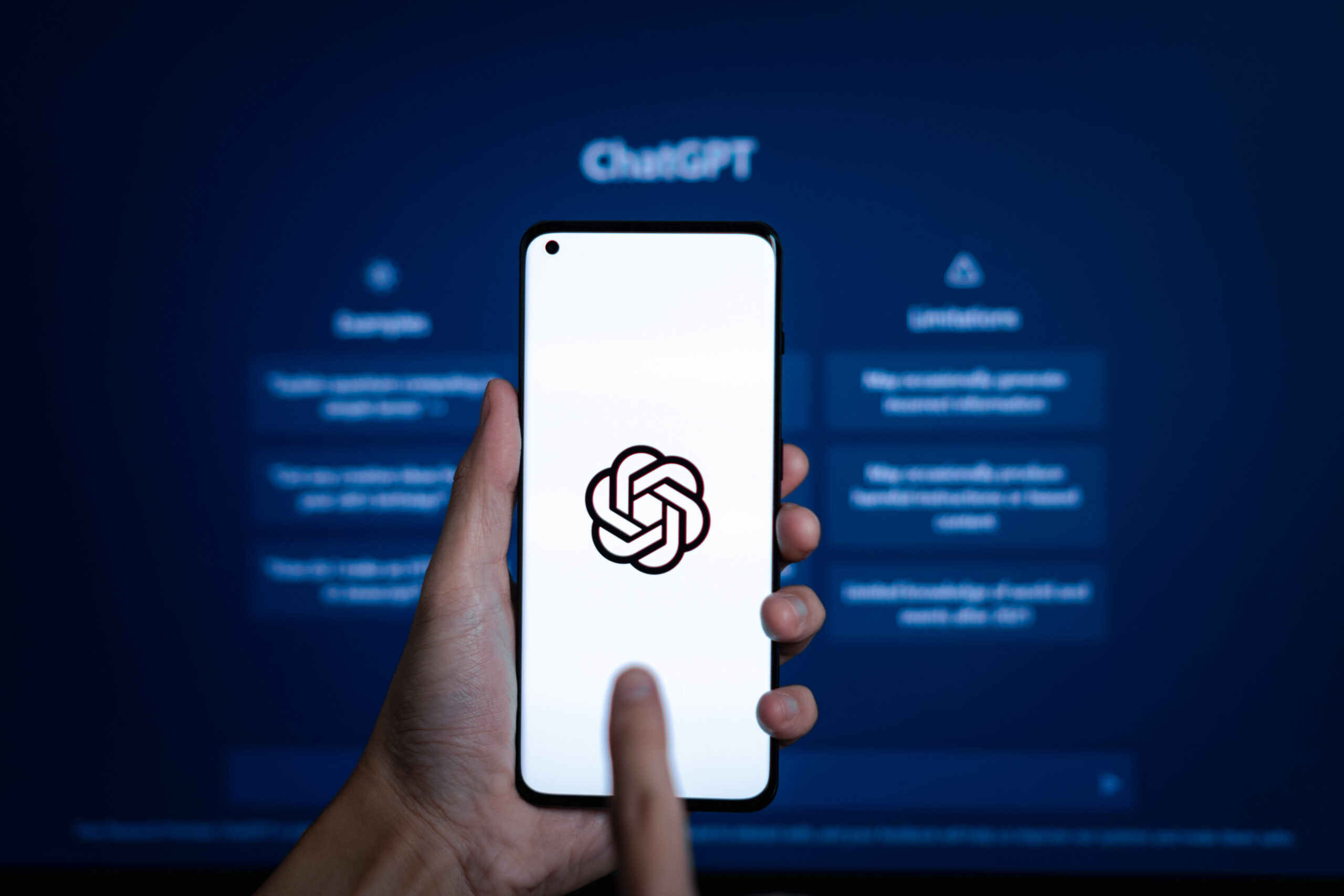 A hand holds a smartphone displaying the ChatGPT logo, with a blurred ChatGPT interface visible on a computer screen in the background, illustrating the search for effective strategies on How To Rank In ChatGPT.
