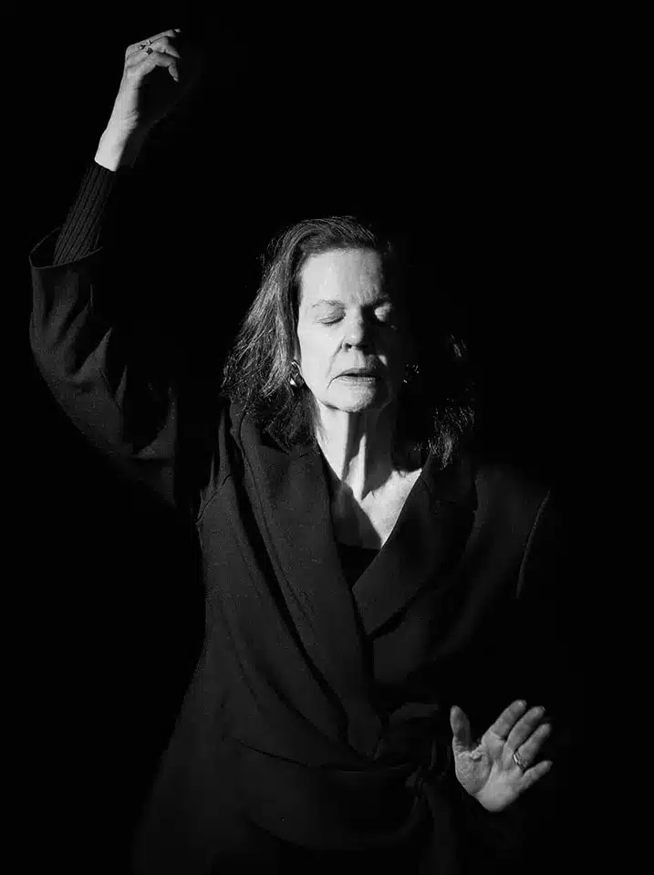 A woman in a dark outfit stands against a black background with her eyes closed, one arm raised and the other hand open, illuminated by a single light source.
