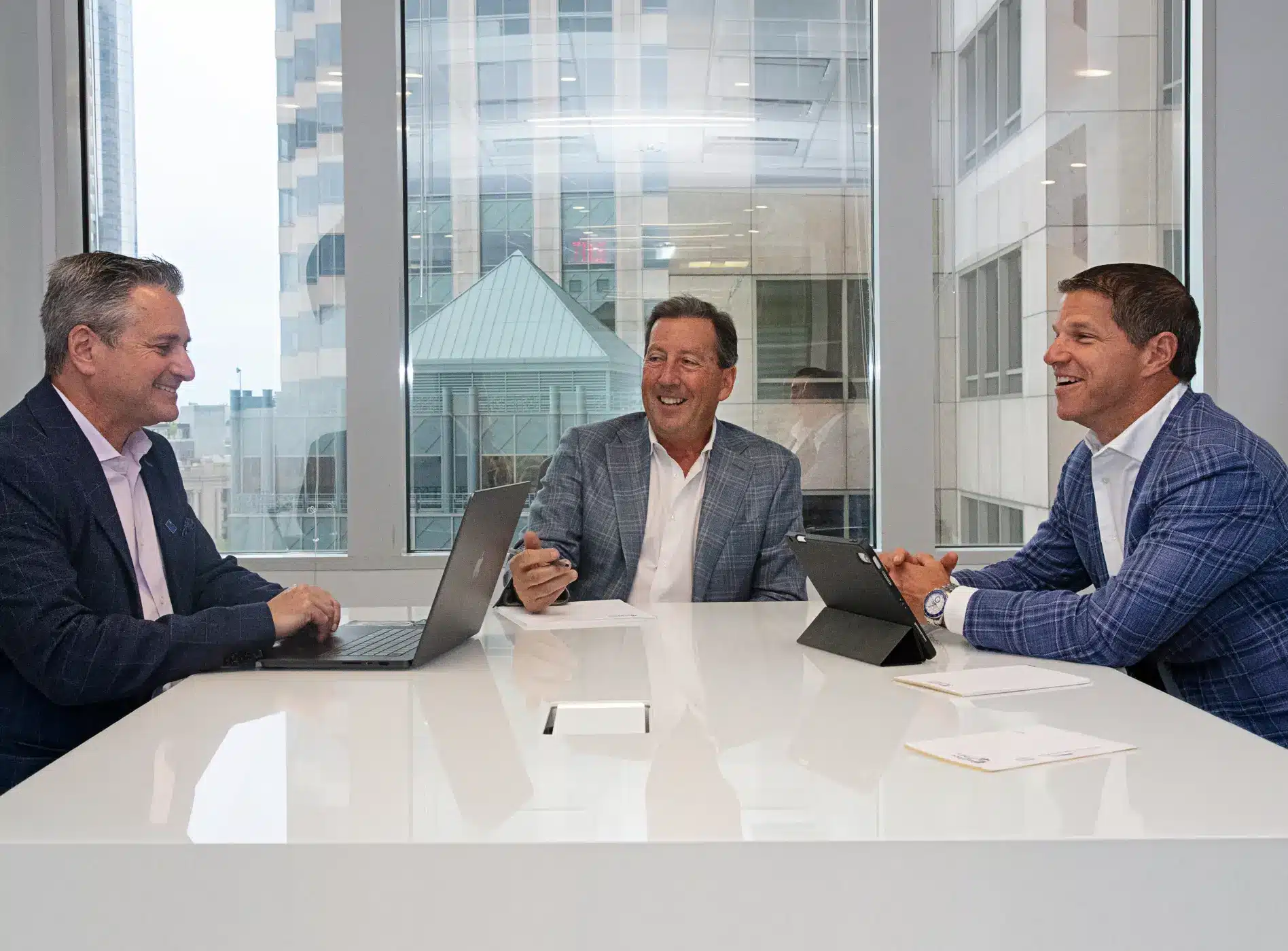 Three men in business attire sit at a white conference table with laptops and papers, having a discussion in a modern office with large windows—an atmosphere reflecting the professionalism of Walter Shuffain.